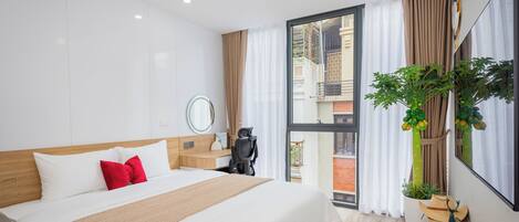 A modern hotel room with a large bed, a desk, a chair, a potted plant, and a view of the outside balcony.