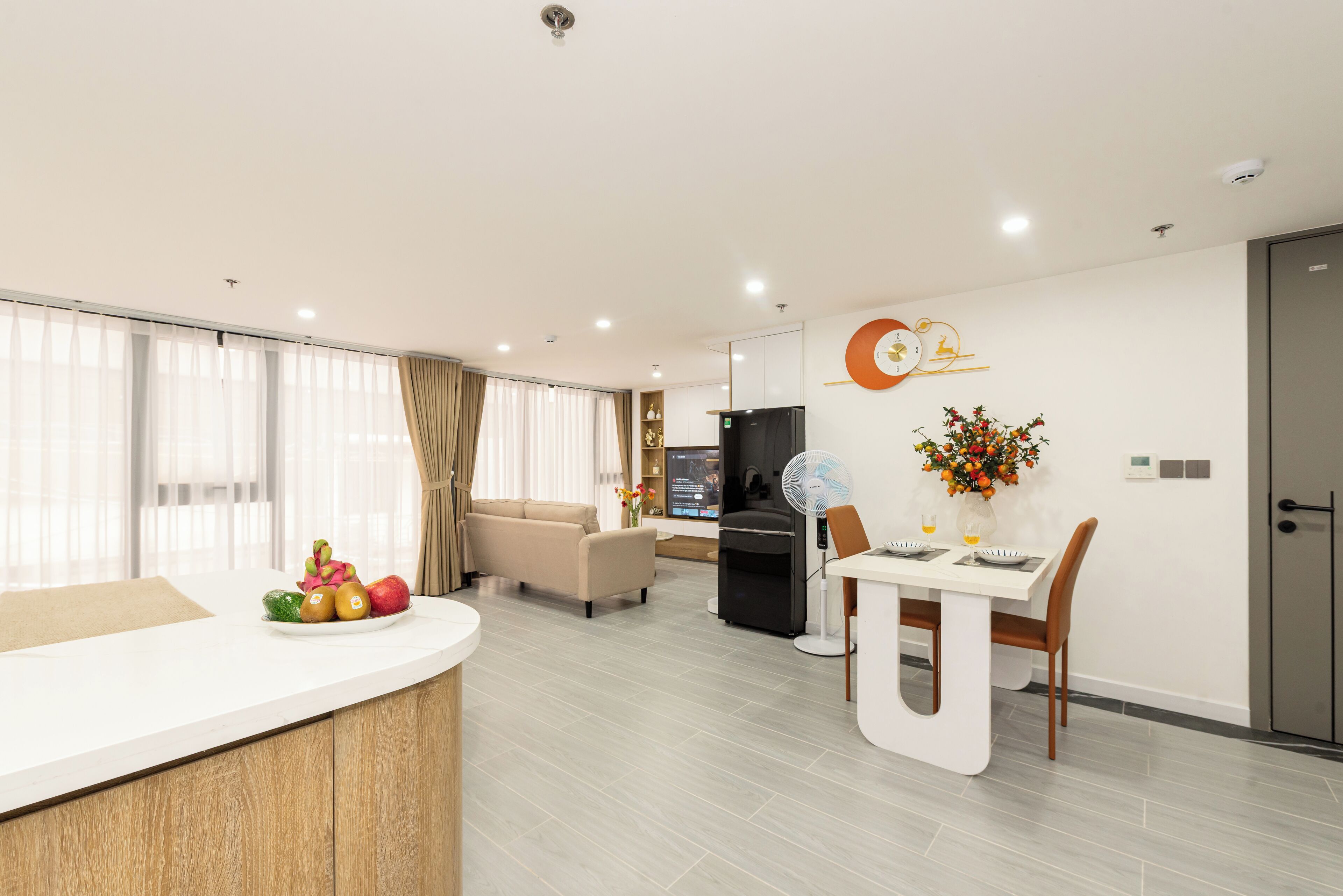 A modern living room with a sofa, a dining table with chairs, a kitchen area with a refrigerator, and a large window with curtains.
