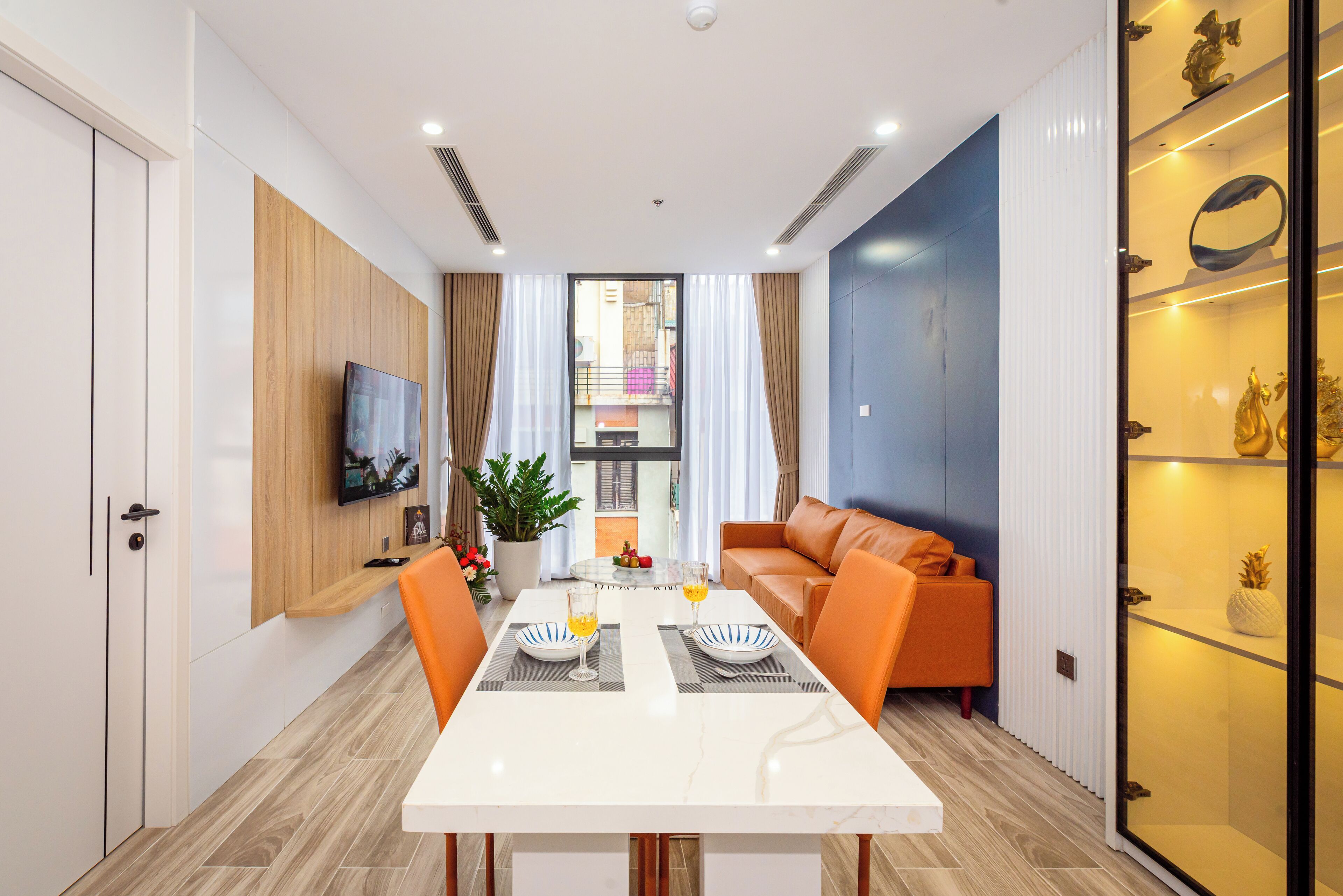 A modern living room with a white dining table, orange sofa, and a TV mounted on the wall.