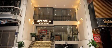 A two-story MYRCELLA APARTMENT building with a staircase leading to the entrance, a covered motorcycle, and a sign indicating the building's name.