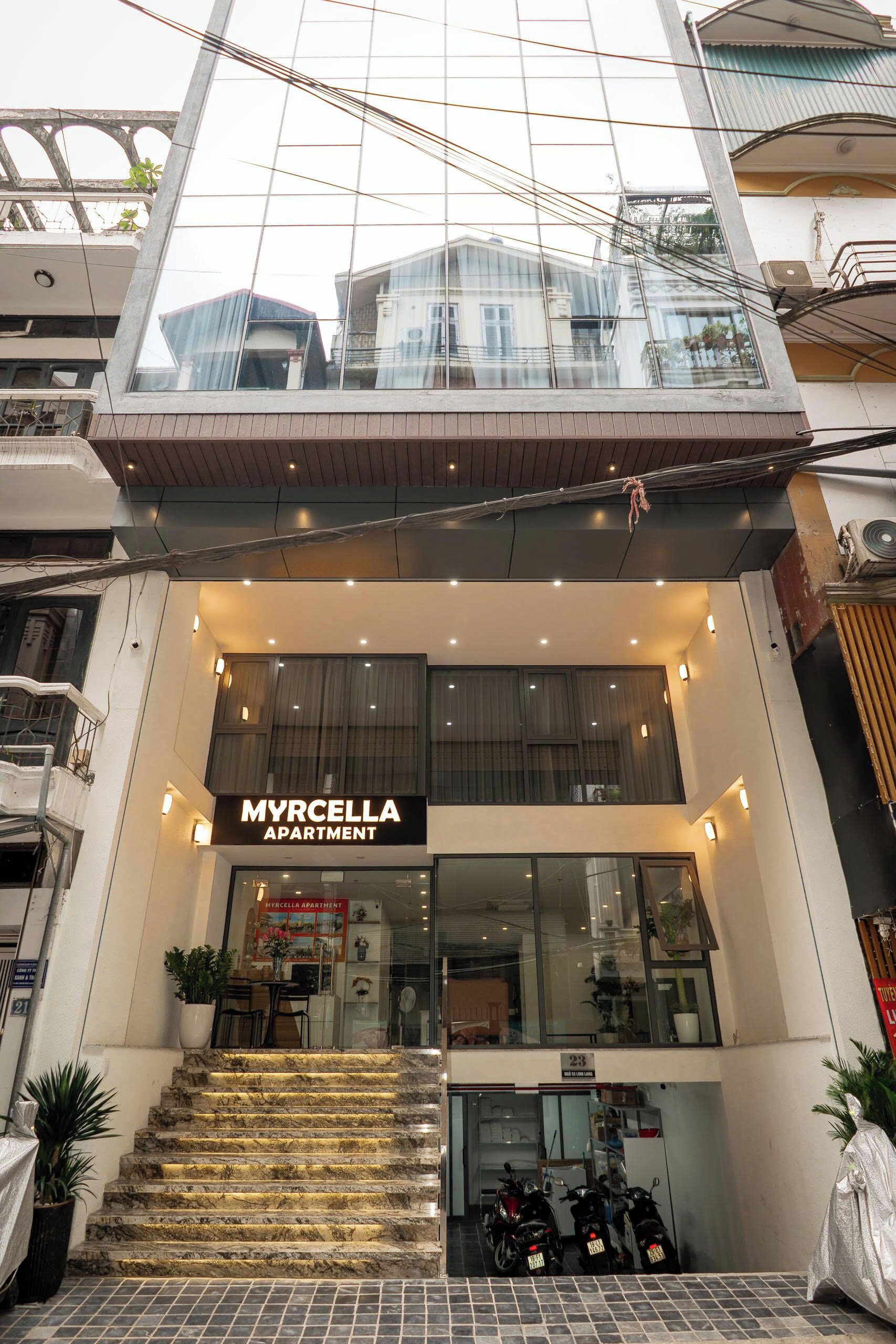 A modern apartment building with a glass facade, a visible entrance, and a sign reading "MYRCELLA APARTMENT".