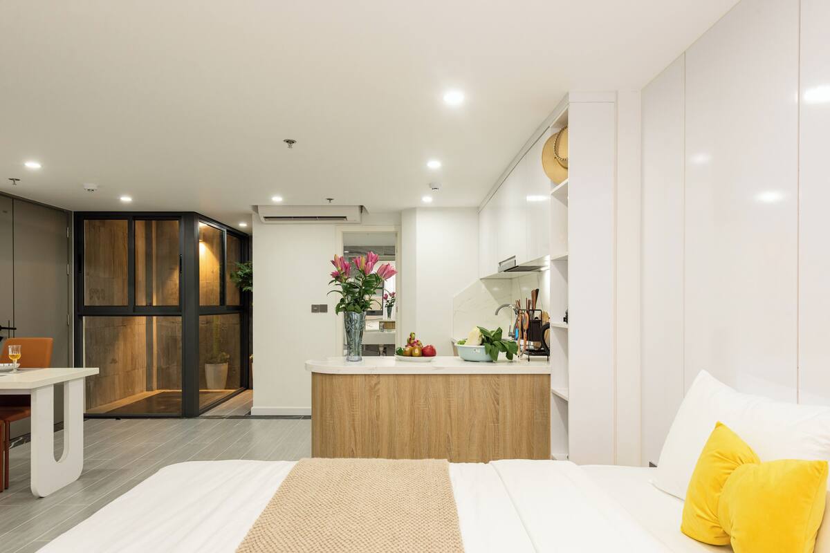 A modern hotel room with a wooden desk, a bed with white and yellow pillows, a kitchen area with a fruit bowl and a vase of flowers, and a large mirror.