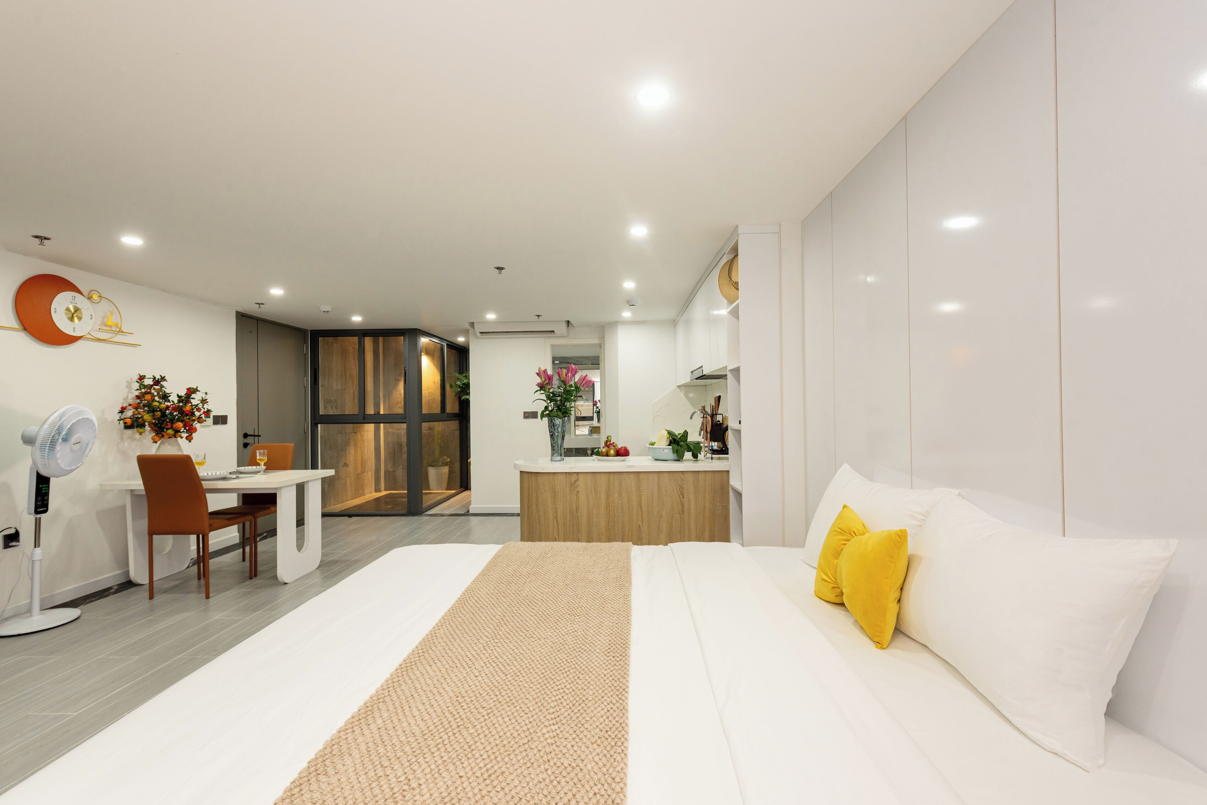 A modern hotel room with a large bed, a desk with a chair, a small kitchenette, and a bathroom visible through an open door.