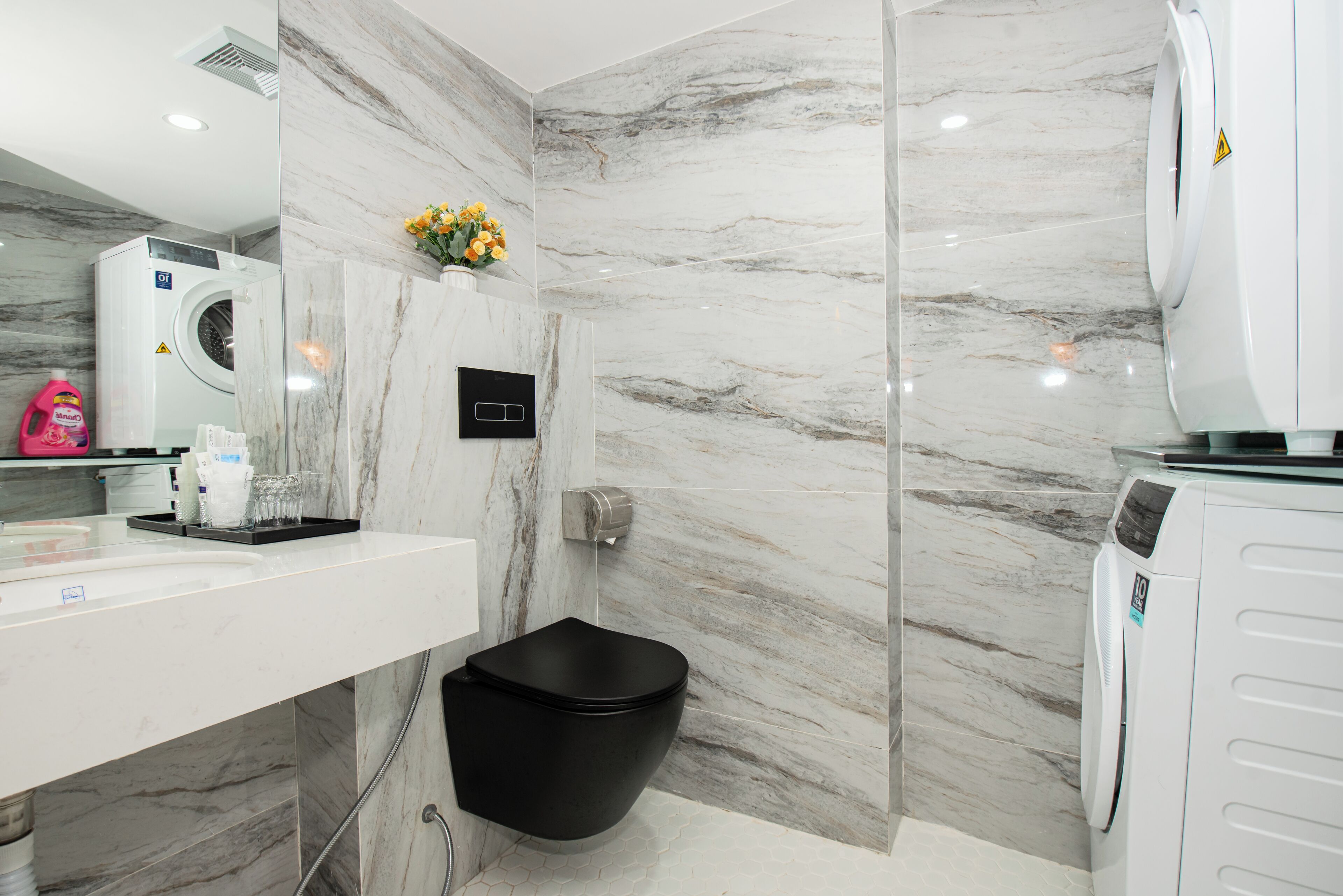 A modern bathroom with a marble wall, a black toilet, a washing machine, and a sink with toiletries.