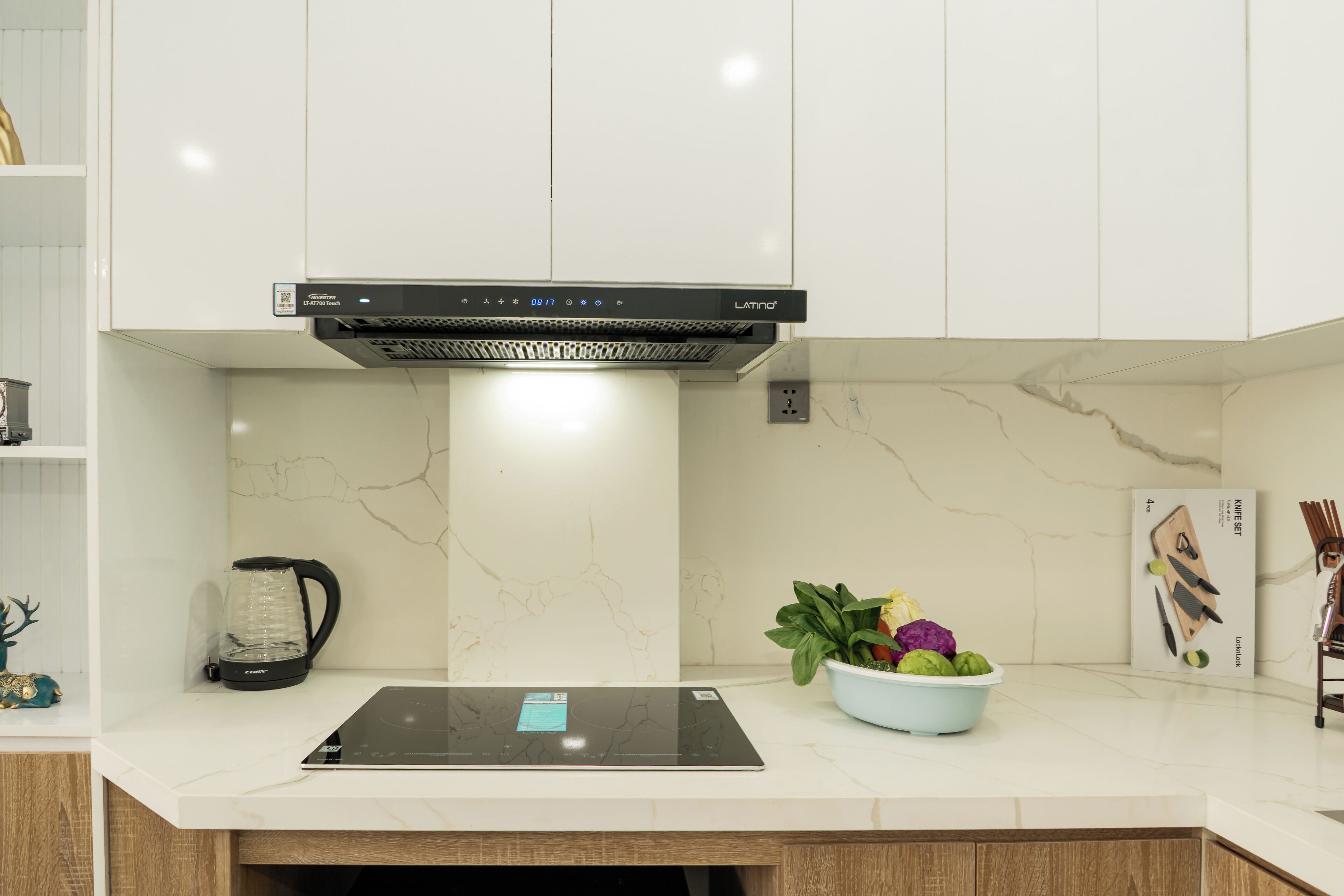 A modern kitchen with white cabinets, a built-in stove, a coffee maker, and a bowl of fresh vegetables.