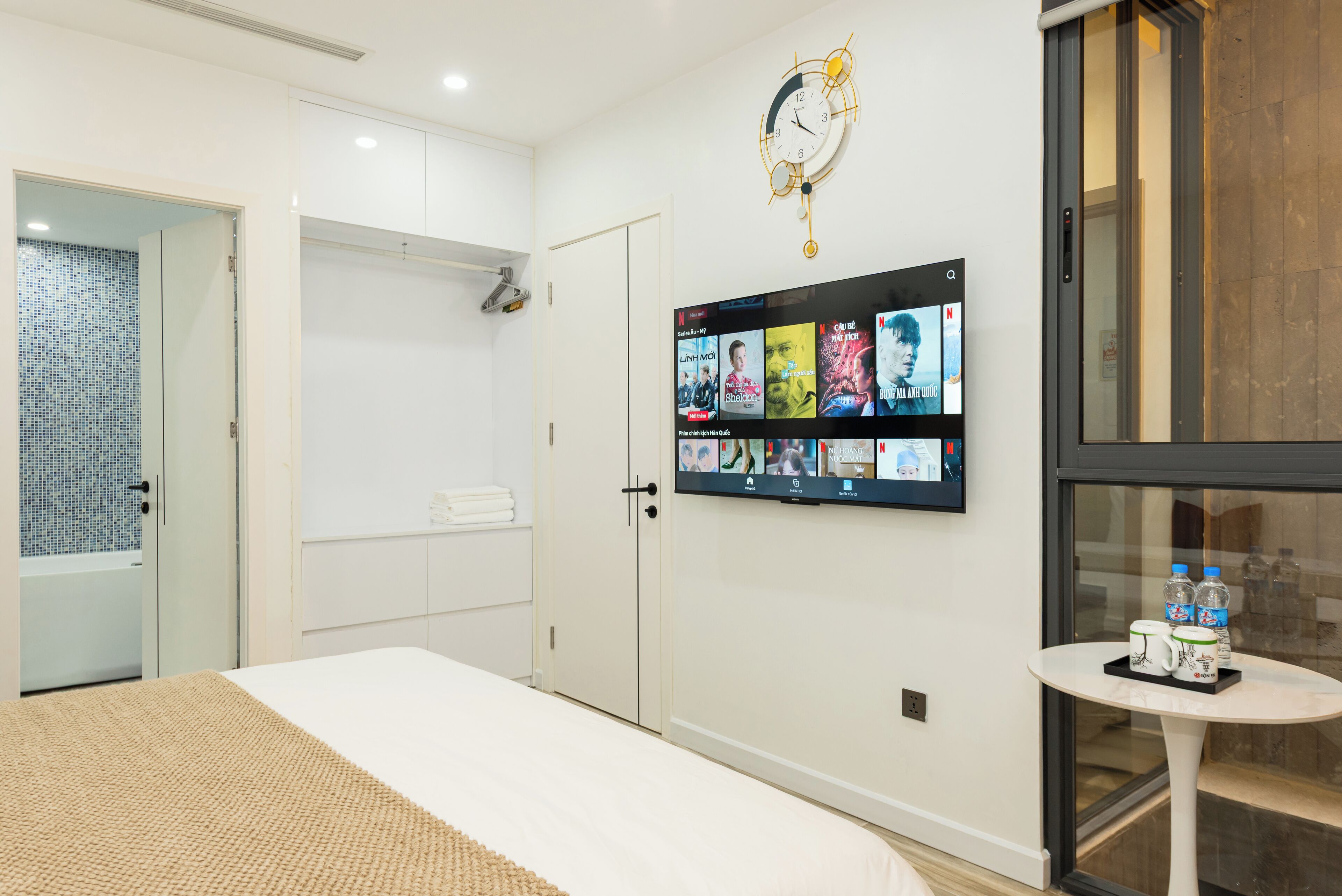 A hotel room with a bed, a TV mounted on the wall displaying various channels, a clock, and a small table with bottles and cups.