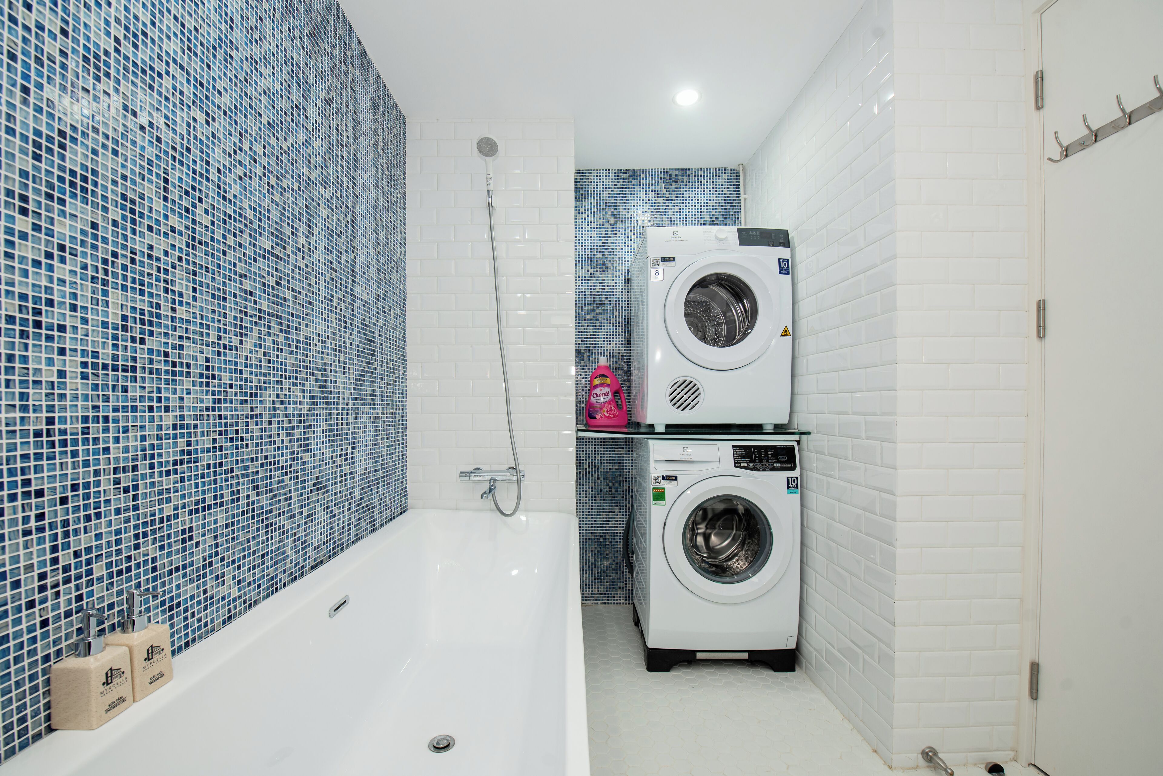 Ein Badezimmer mit einer weißen Badewanne, zwei übereinander gestapelten Waschmaschinen und blauen Mosaikfliesen an den Wänden.