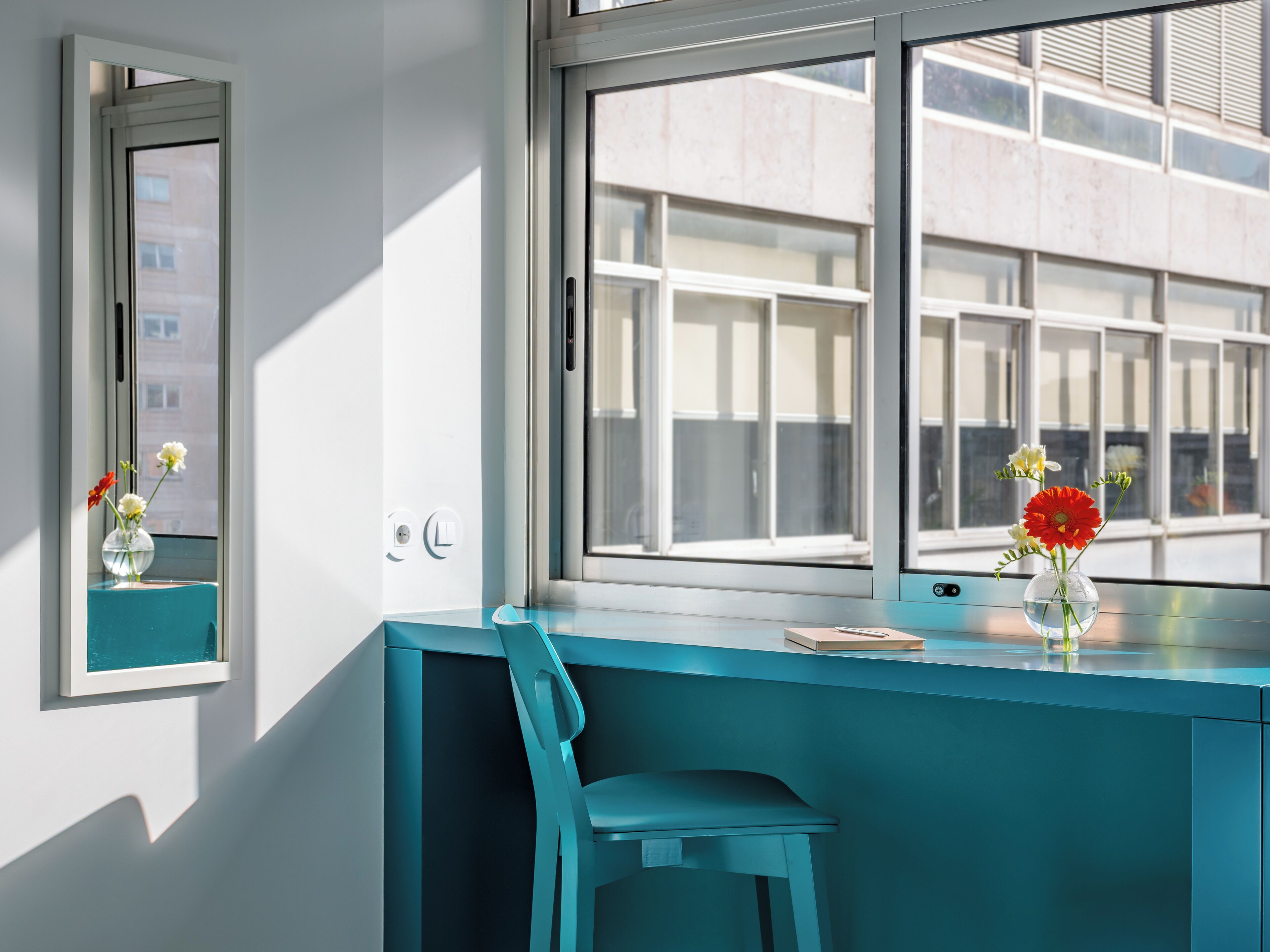 Une pièce moderne avec un bureau de couleur turquoise, une chaise, une fenêtre donnant sur des immeubles et un vase rempli de fleurs posé sur le rebord de la fenêtre.