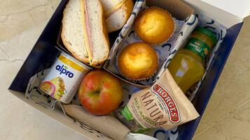 A lunch box containing sandwiches, muffins, a bottle of juice, a jar of peanut butter, a carton of almond milk, and an apple.