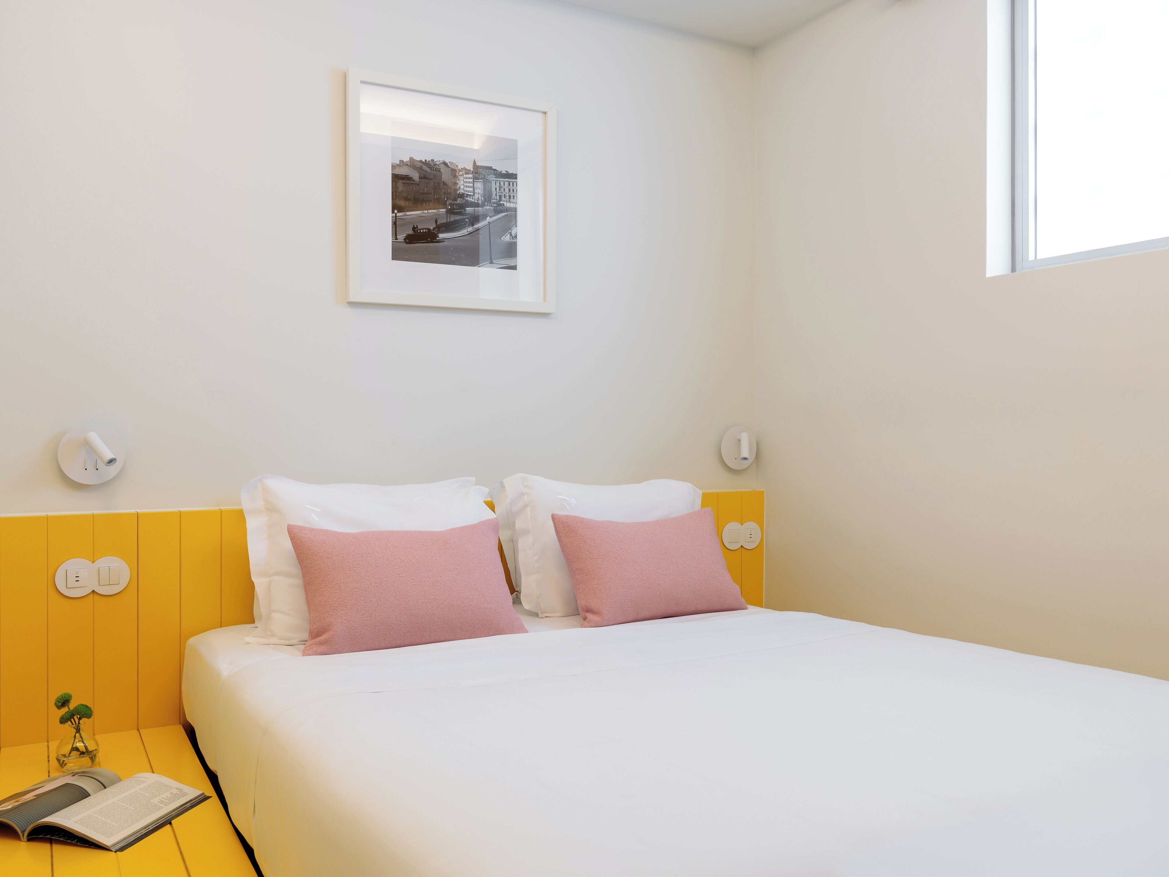 A neatly arranged hotel room with a bed, pillows, a bedside table with a book, and a wall-mounted picture.