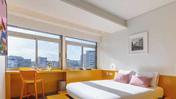 A modern hotel room with a bed, a desk, and a chair. There are large windows with a city view.