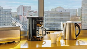 A kitchen with a coffee maker, two cups, and a kettle on a yellow countertop, with a city view through a window.