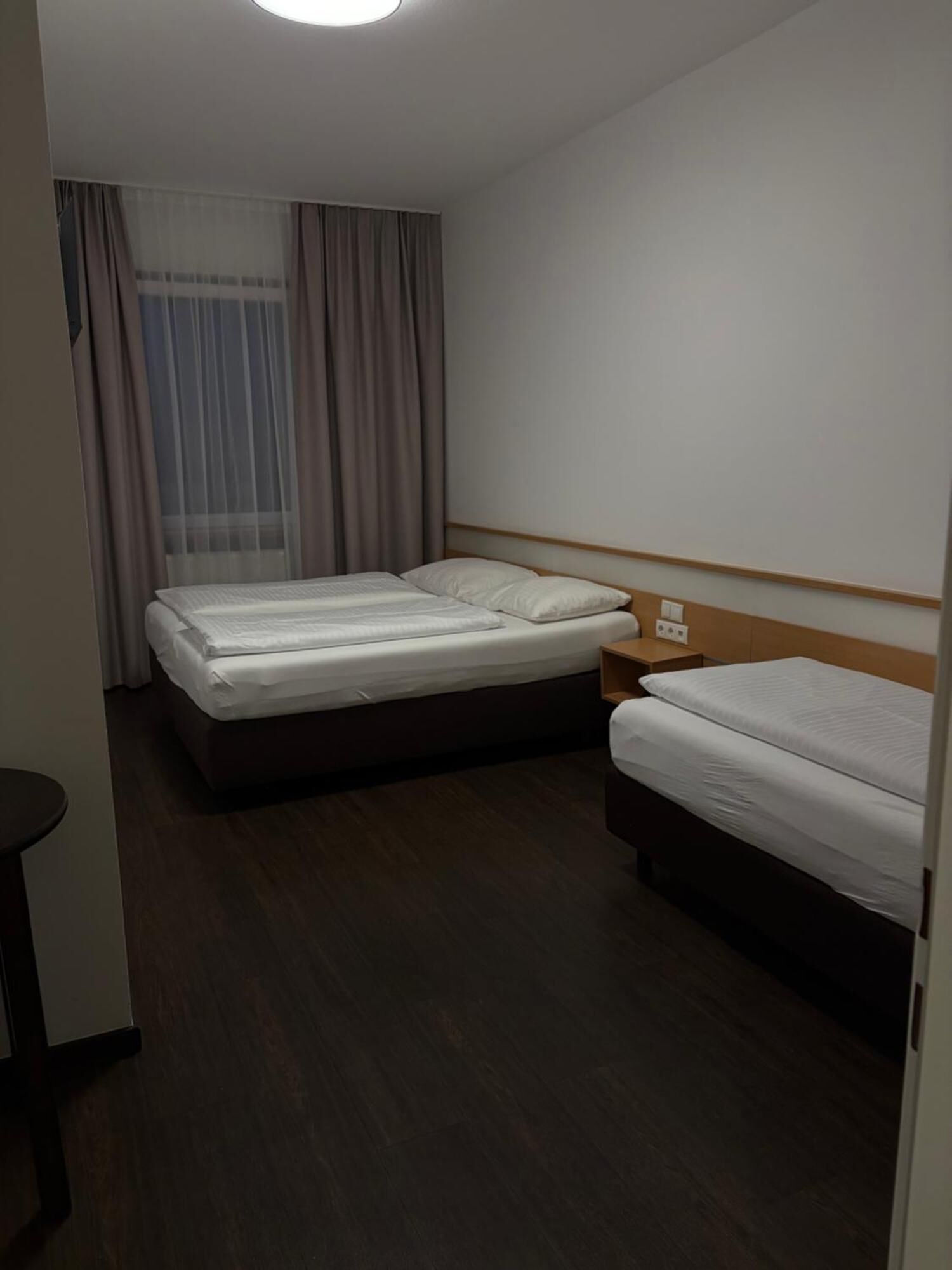 A hotel room with two beds, a wooden nightstand, a small table, and a window with curtains.
