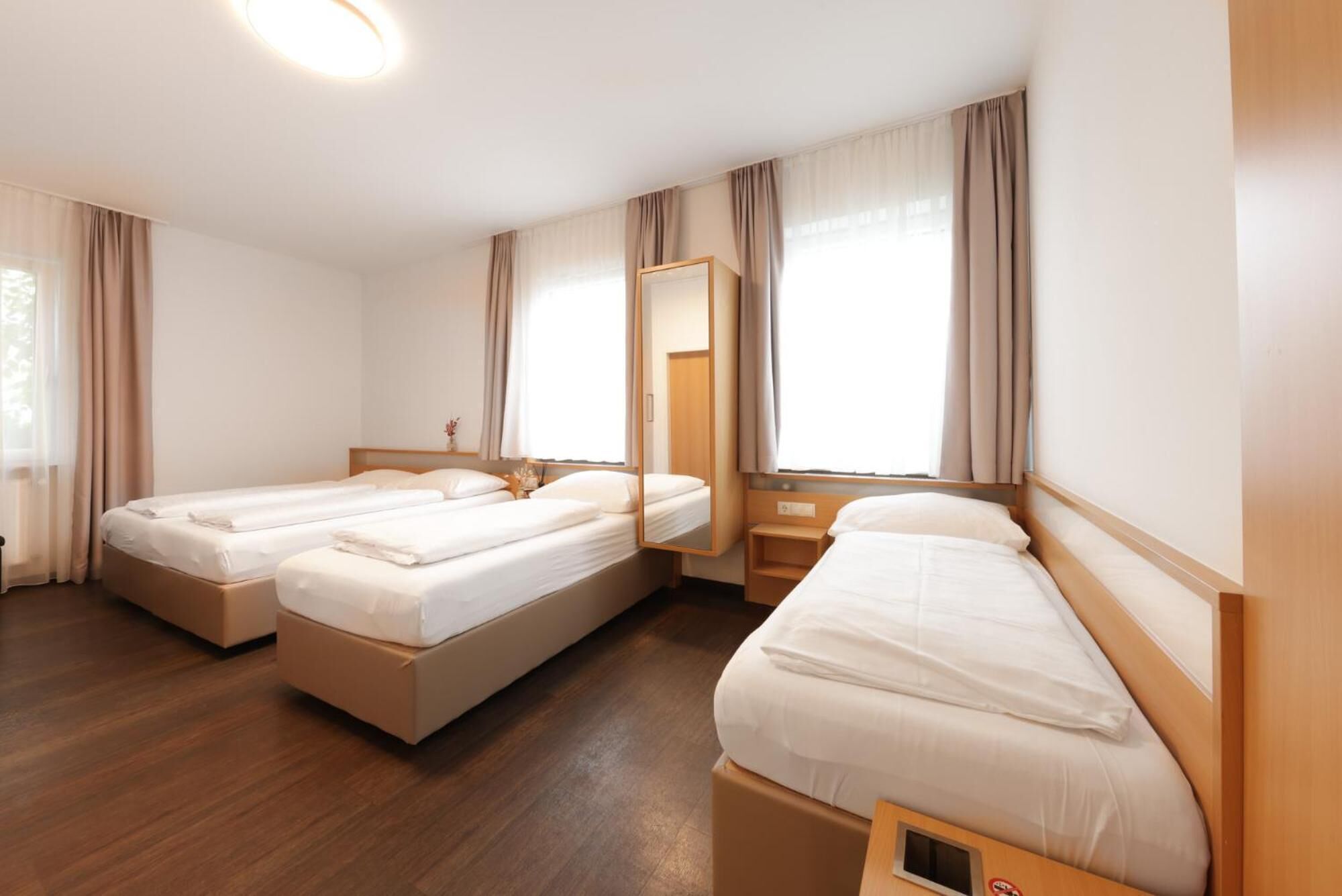 Habitación de hotel con dos camas individuales, cabecera de madera, mesita de noche, espejo y ventana con cortinas.