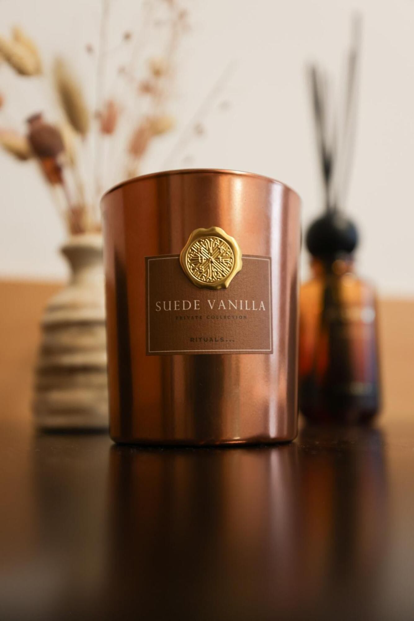 A copper-colored candle labeled "SUEDE VANILLA" with a gold emblem, placed on a wooden surface.