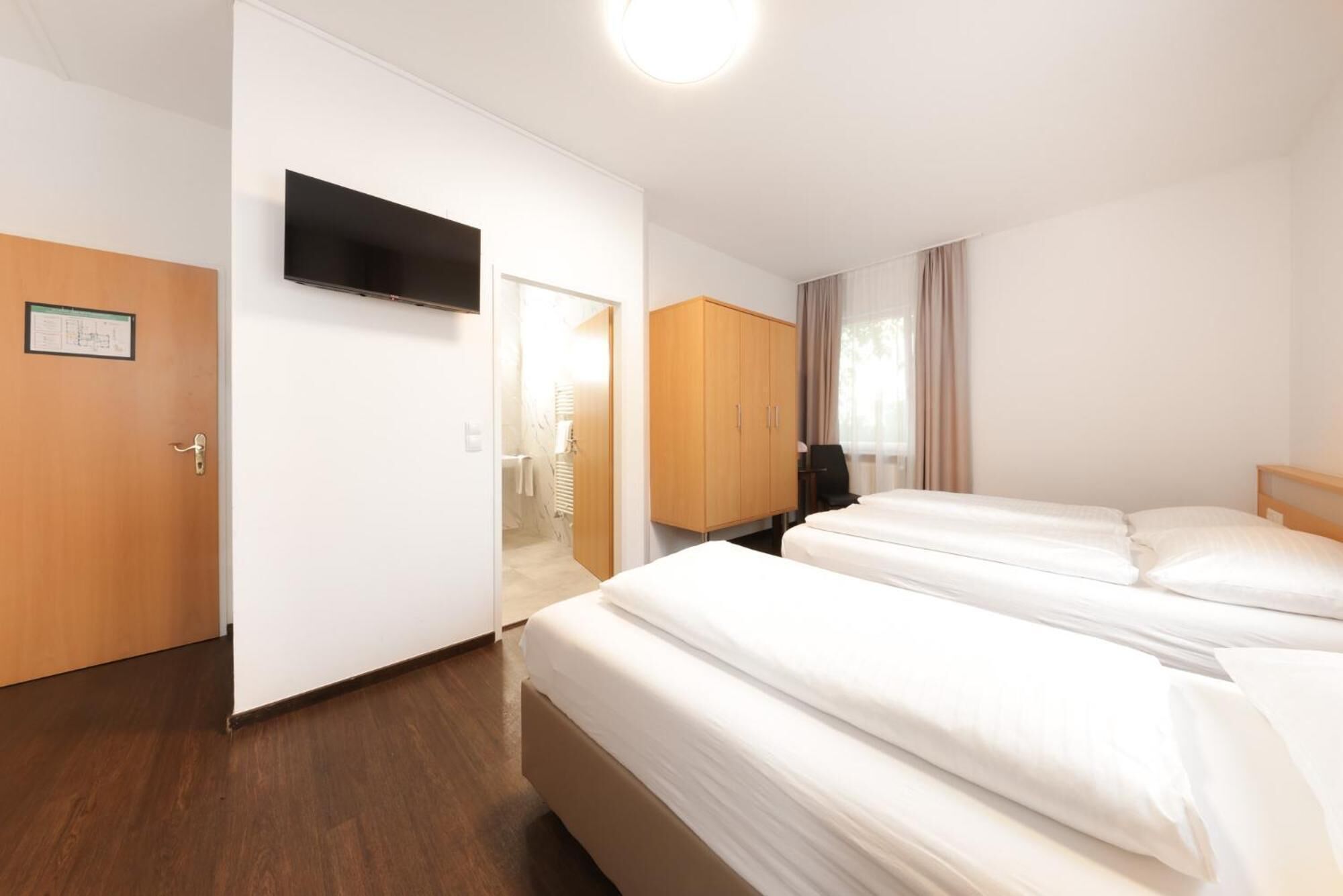 A hotel room with two beds, a TV mounted on the wall, a wooden wardrobe, and a desk with a chair.