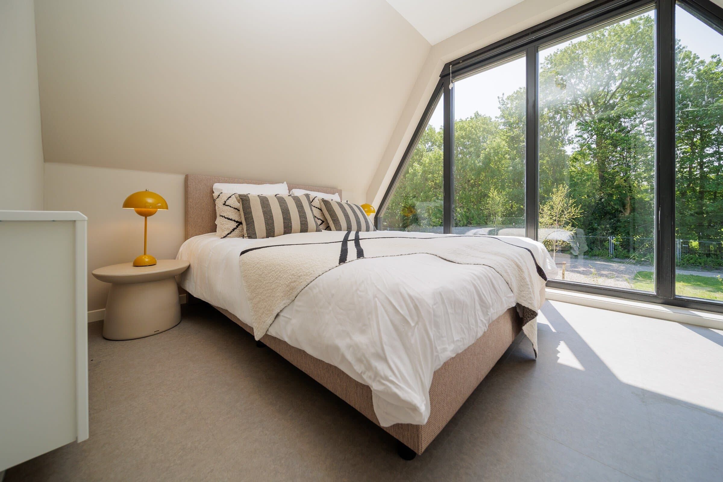 A bedroom with a bed, bedside table, lamp, and large window with a view of greenery.