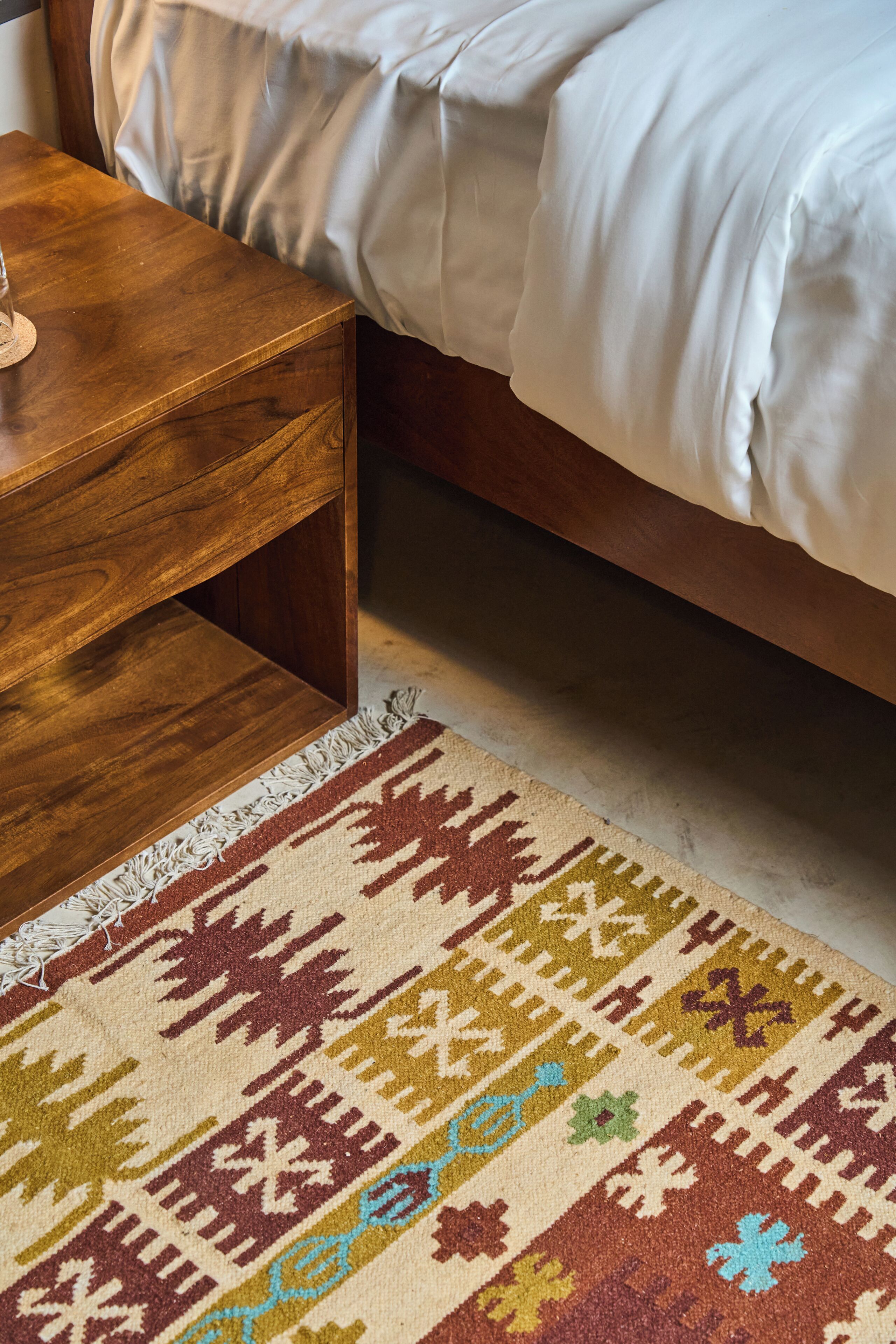 Une table de chevet en bois, un tapis à motifs et un lit avec une literie blanche.