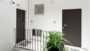 Un bâtiment doté de deux portes sombres, d’une petite fenêtre avec des stores, d’un pot de fleurs et d’une rambarde en métal.