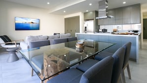Een moderne woonkamer met een glazen salontafel, grijze banken en een grote flatscreen televisie aan de muur.