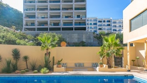 Una piscina con una tumbona y palmeras, rodeada por un muro bajo y un edificio al fondo.