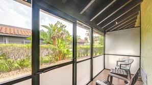 A covered balcony with a view of a garden, a chair, and a table.