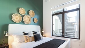 A neatly made bed with a white headboard, a black throw blanket, and several pillows. There are woven wall decorations above the bed and a large window with a view of a staircase. - New Build | Sleeps 12, 2 Condos in Scenic Bluffside, Minutes to Downtown (Nashville)