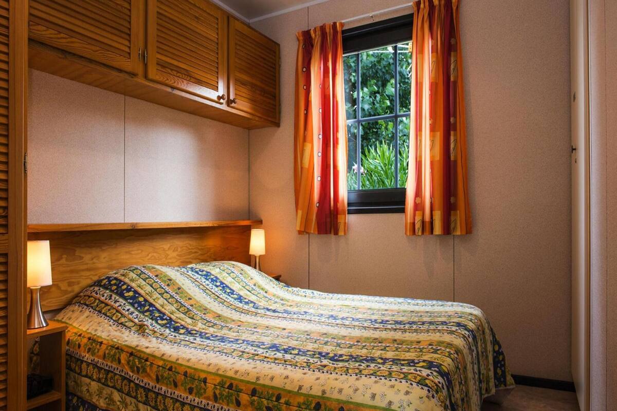 A bedroom with a wooden headboard, a patterned bedspread, two bedside lamps, and a window with curtains.