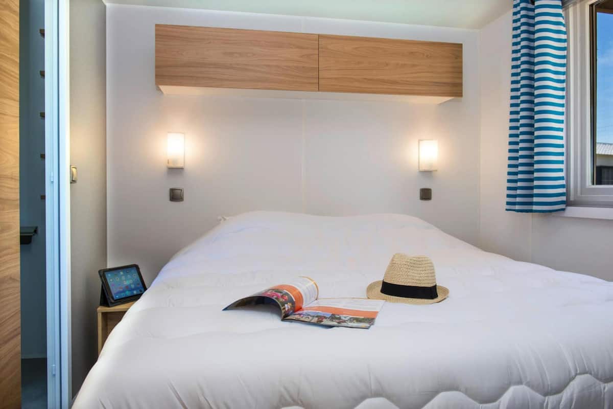 A neatly made bed with a magazine and a straw hat on it, a bedside table with a tablet, and two wall-mounted lights.