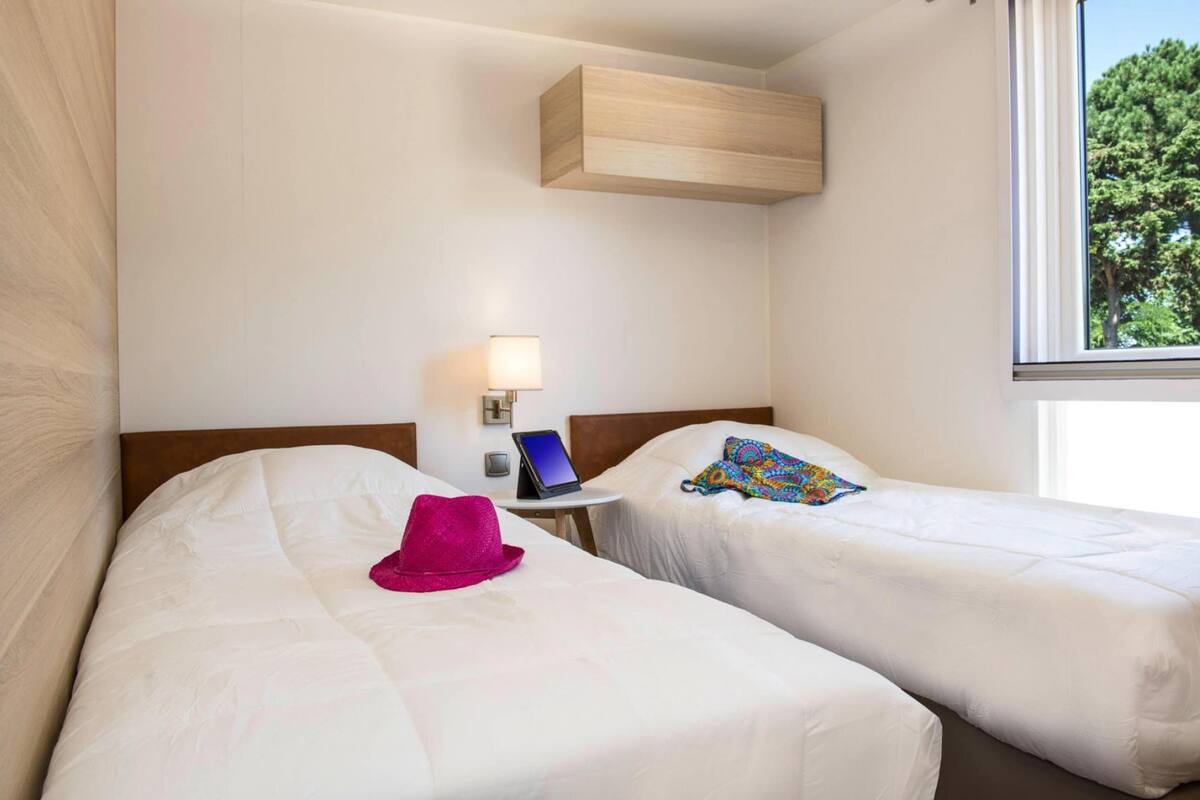 Ein Hotelzimmer mit zwei Betten, einem Nachttisch mit Lampe, einem Tablet und einem Fenster mit Blick auf B äume.