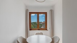 Une pièce avec une table ronde et deux chaises, une fenêtre avec des rideaux et une vue sur la verdure et les bâtiments.