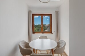 Quarto com uma mesa redonda e duas cadeiras, janela com cortinas e vista para áreas verdes e edifícios.