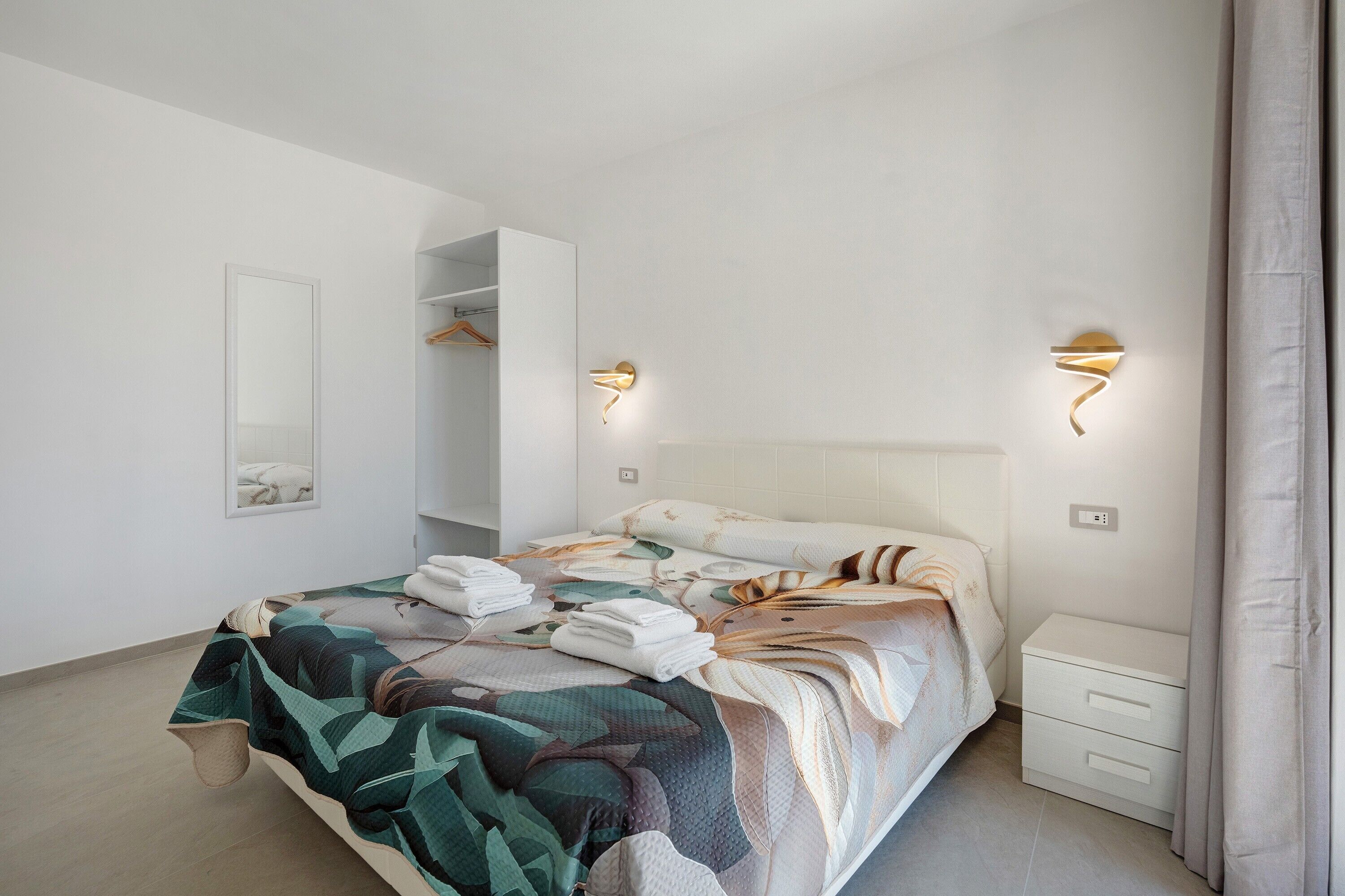 A bedroom with a bed, a bedside table, a mirror, and wall-mounted lights.