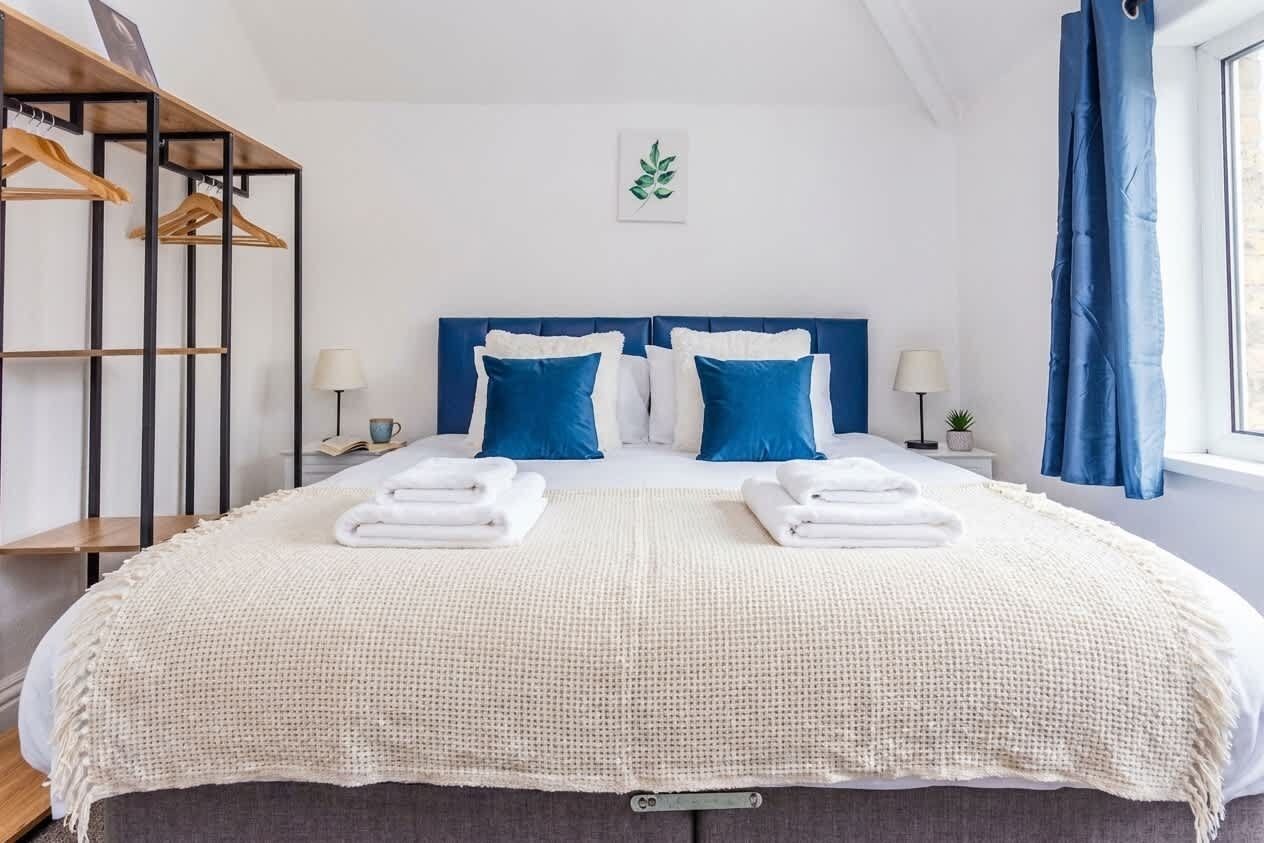 Un lit double bien fait, aux couleurs blanche et bleue, une étagère en bois avec des vêtements suspendus et une petite table de chevet avec une lampe.