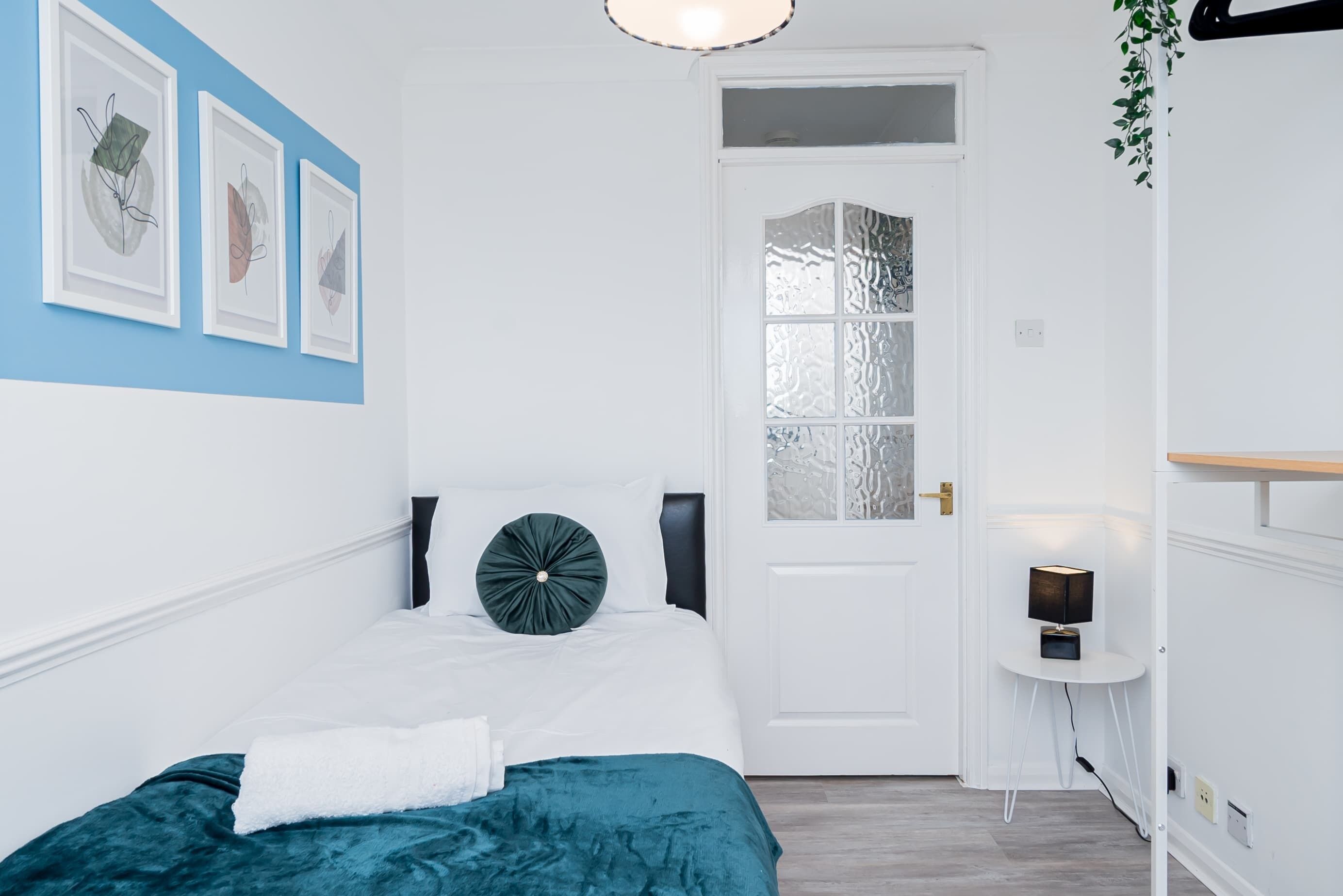 A bedroom with a bed, a white door, a small table with a lamp, and wall art.
