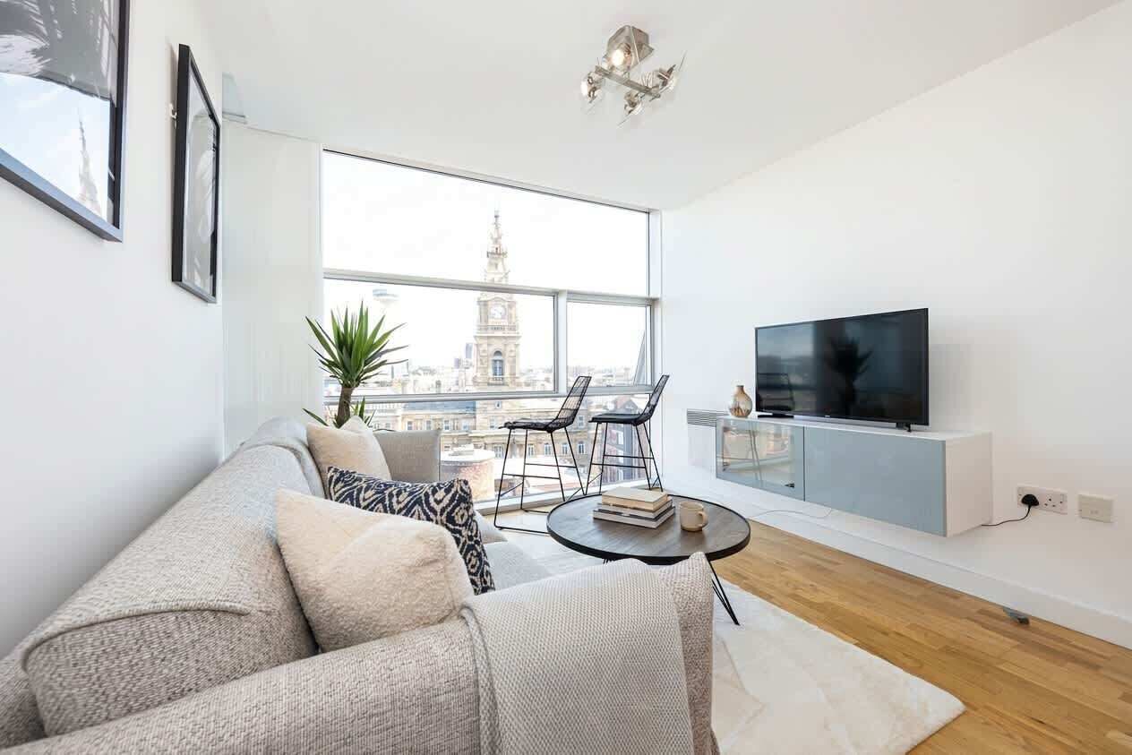 A modern living room with a sofa, a coffee table, a TV, and a large window offering a city view.