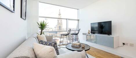 A modern living room with a sofa, a coffee table, a TV, and a large window offering a city view.