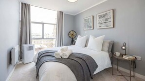 A bedroom with a large bed, a standing fan, a small table with a lamp, and artwork on the wall. - Stunning Skyline 1BR Liverpool Apartment w Parking (Liverpool)