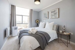 Une chambre à coucher avec un grand lit, un ventilateur sur pied, une petite table avec une lampe et des œuvres d’art accrochées au mur.