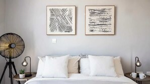 Een slaapkamer met een bed, twee nachtkastjes, een vloerlamp, een staande ventilator en twee kunstwerken in lijsten aan de muur.