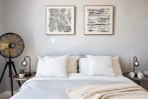 Un dormitorio con una cama, dos mesitas de noche, una lámpara de pie, un ventilador y dos cuadros enmarcados en la pared.
