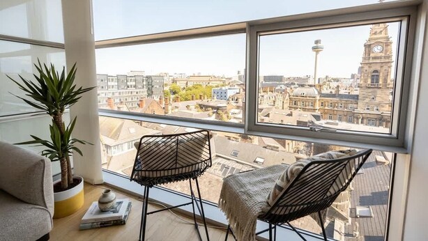 A room with a large window overlooking a cityscape, featuring a potted plant, two chairs, and a sofa. - Stunning Skyline 1BR Liverpool Apartment w Parking (Liverpool)