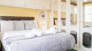 A neatly made bed with a grey headboard and pillows, a small shelf with a vase, and a wardrobe with mirrored doors and a hanging light.