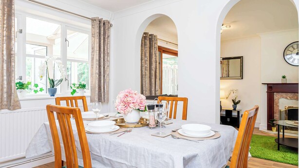 A dining room with a table set for six, surrounded by wooden chairs, white tablecloth, and floral centerpieces.