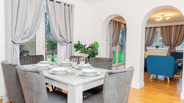 Un coin repas avec une table blanche dressée pour quatre personnes, des chaises à revêtement gris et des rideaux gris clair.