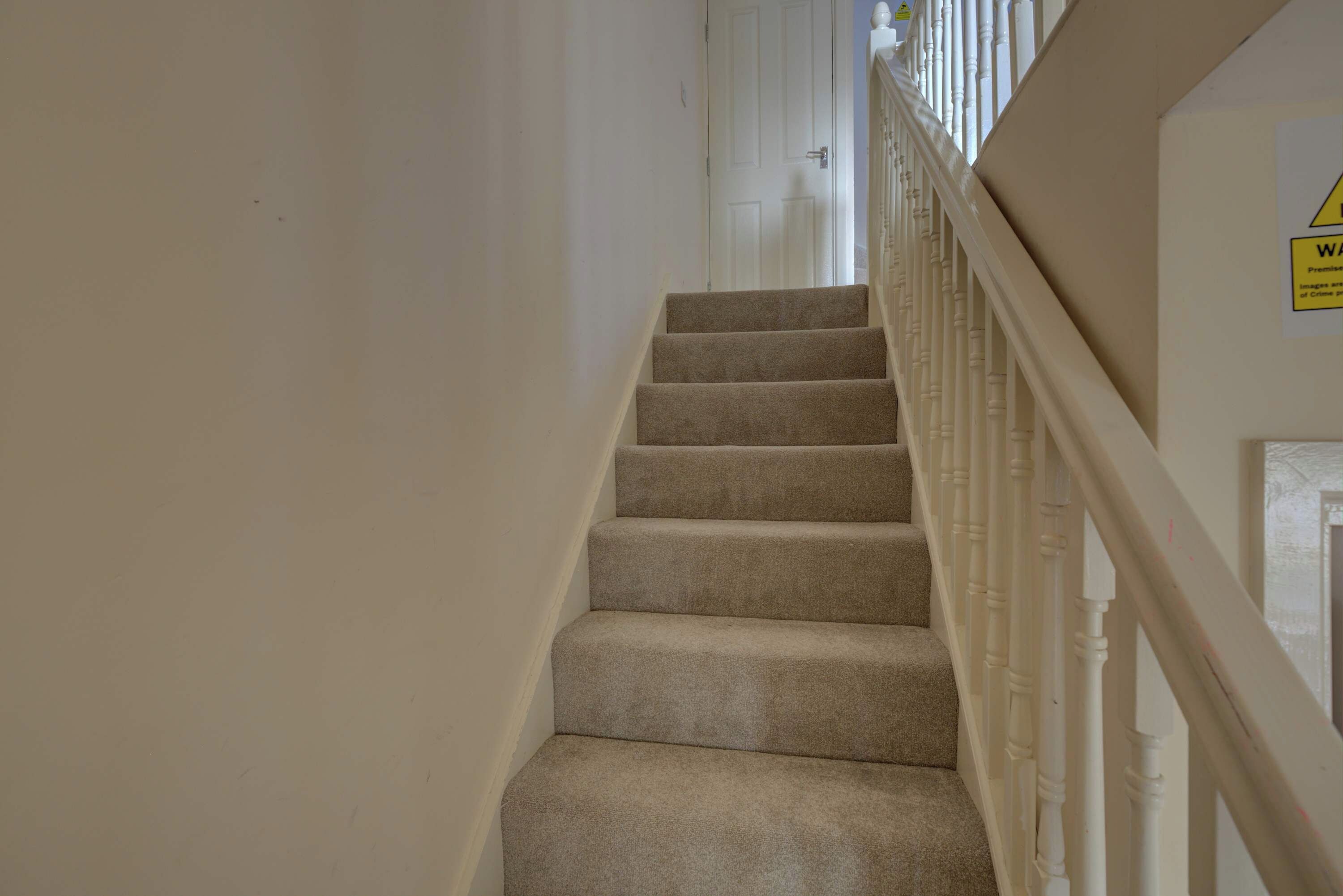 Un escalier recouvert de moquette, avec des rampes blanches et une porte en haut.