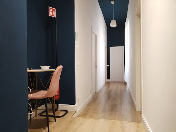 Un pasillo estrecho con suelo de madera, una puerta blanca al final y una mesa con sillas a la izquierda.
