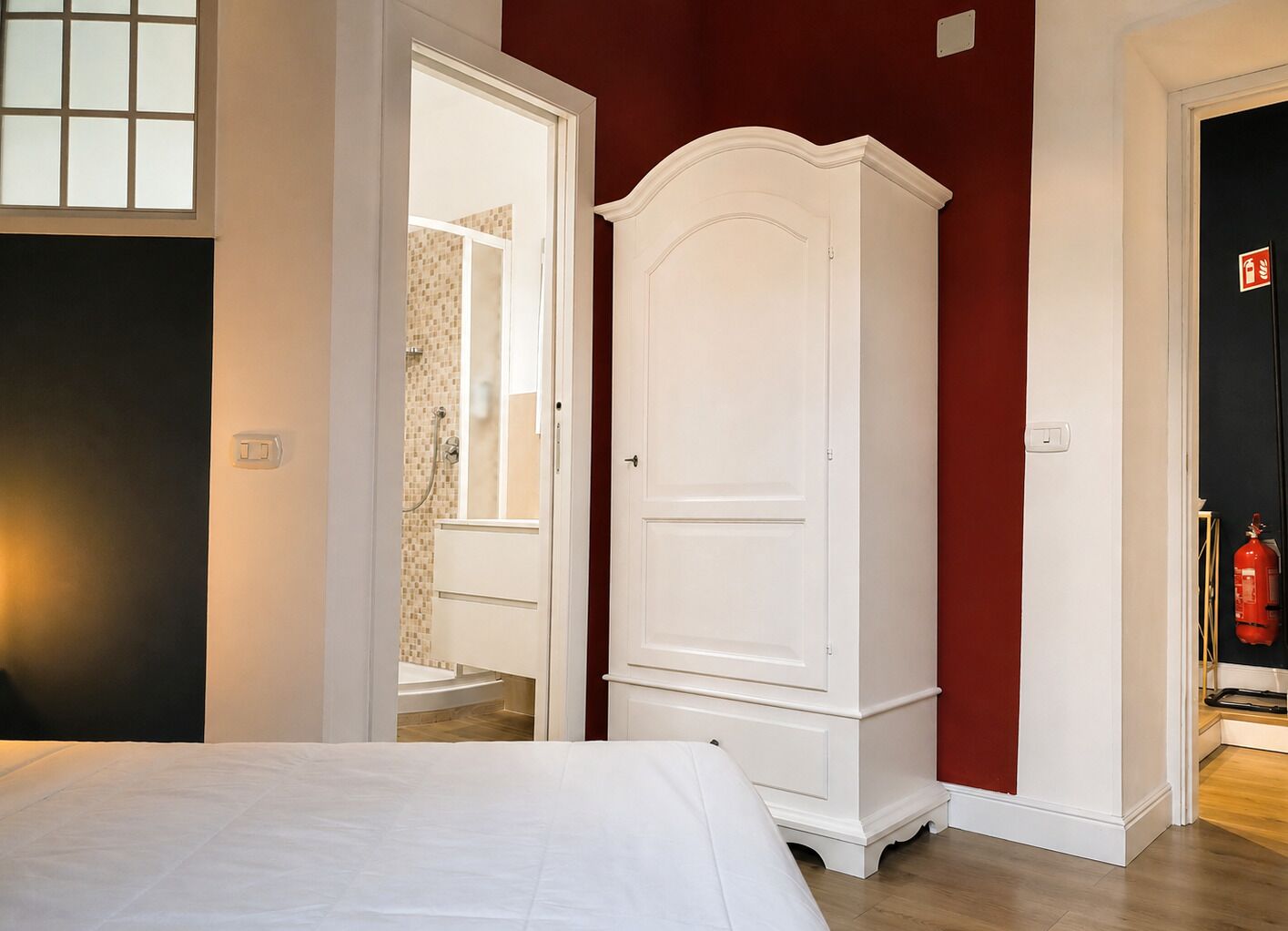 A bedroom with a bed, a white wardrobe, a red wall, and a bathroom with a shower and a toilet visible through an open door.