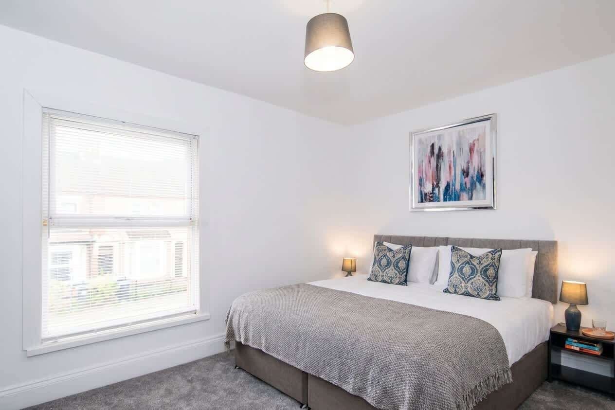 Un dormitorio con cama, lámparas de noche, ventana con persianas, luz de techo y una obra de arte enmarcada en la pared.