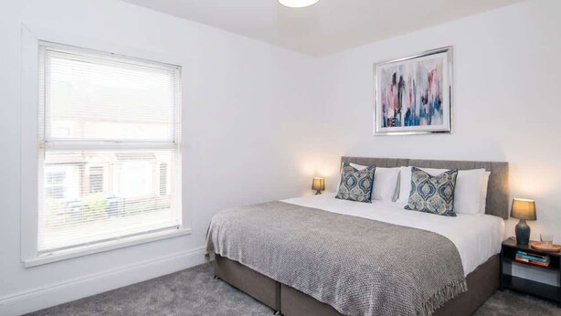 A bedroom with a bed, bedside lamps, a window with blinds, a ceiling light, and a framed artwork on the wall. - Modern 3BR Home with Free Parking & Fast Wi-Fi (Rugby)